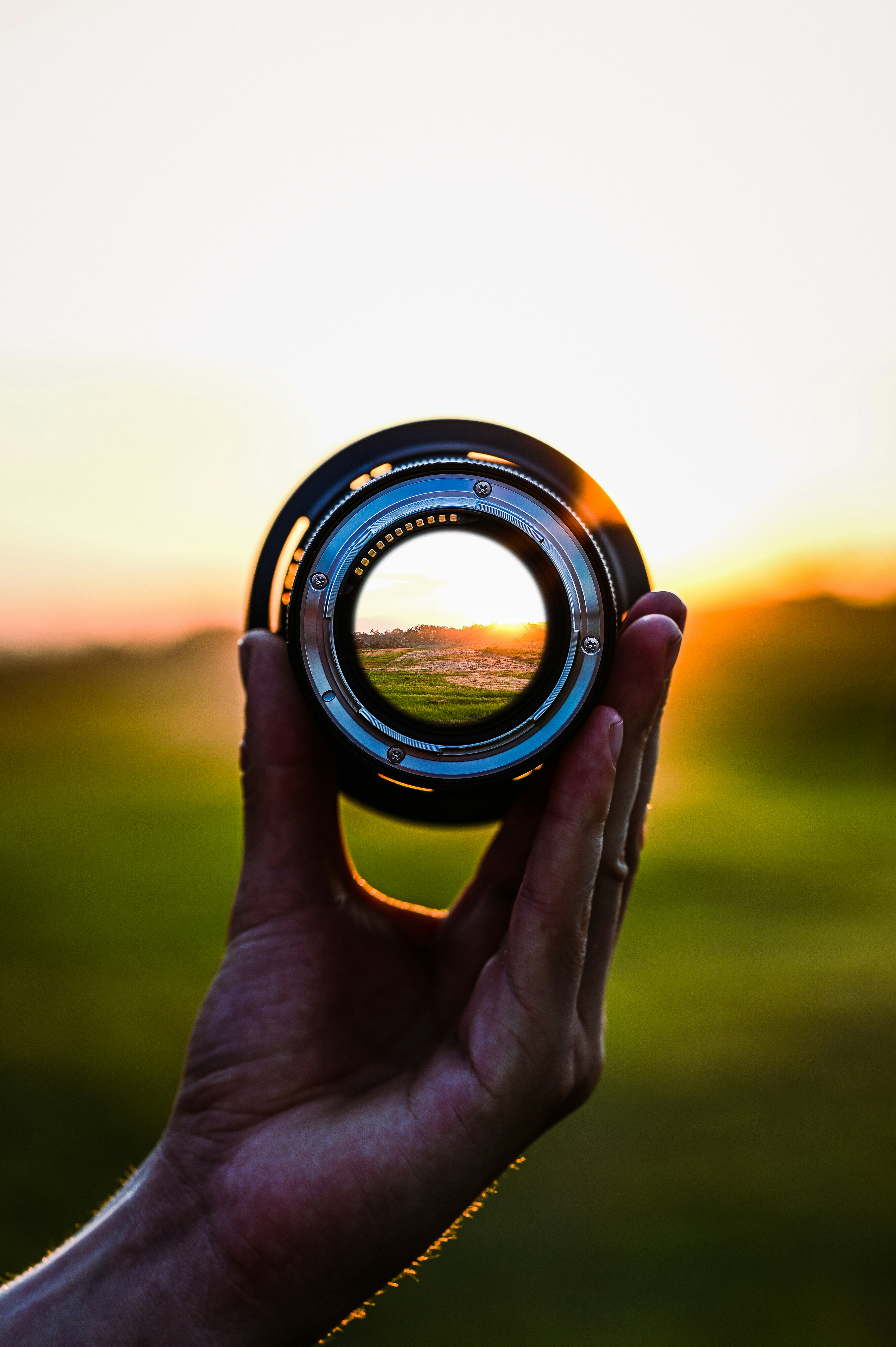 A hand holding a camera lens with a warm sunset visible through the glass, creating a beautiful bokeh effect with golden and amber tones